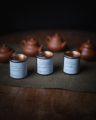 Three tea tins with white labels and metallic lids are arranged on a dark cloth over a wooden surface, with blurred terracotta teapots in the background.