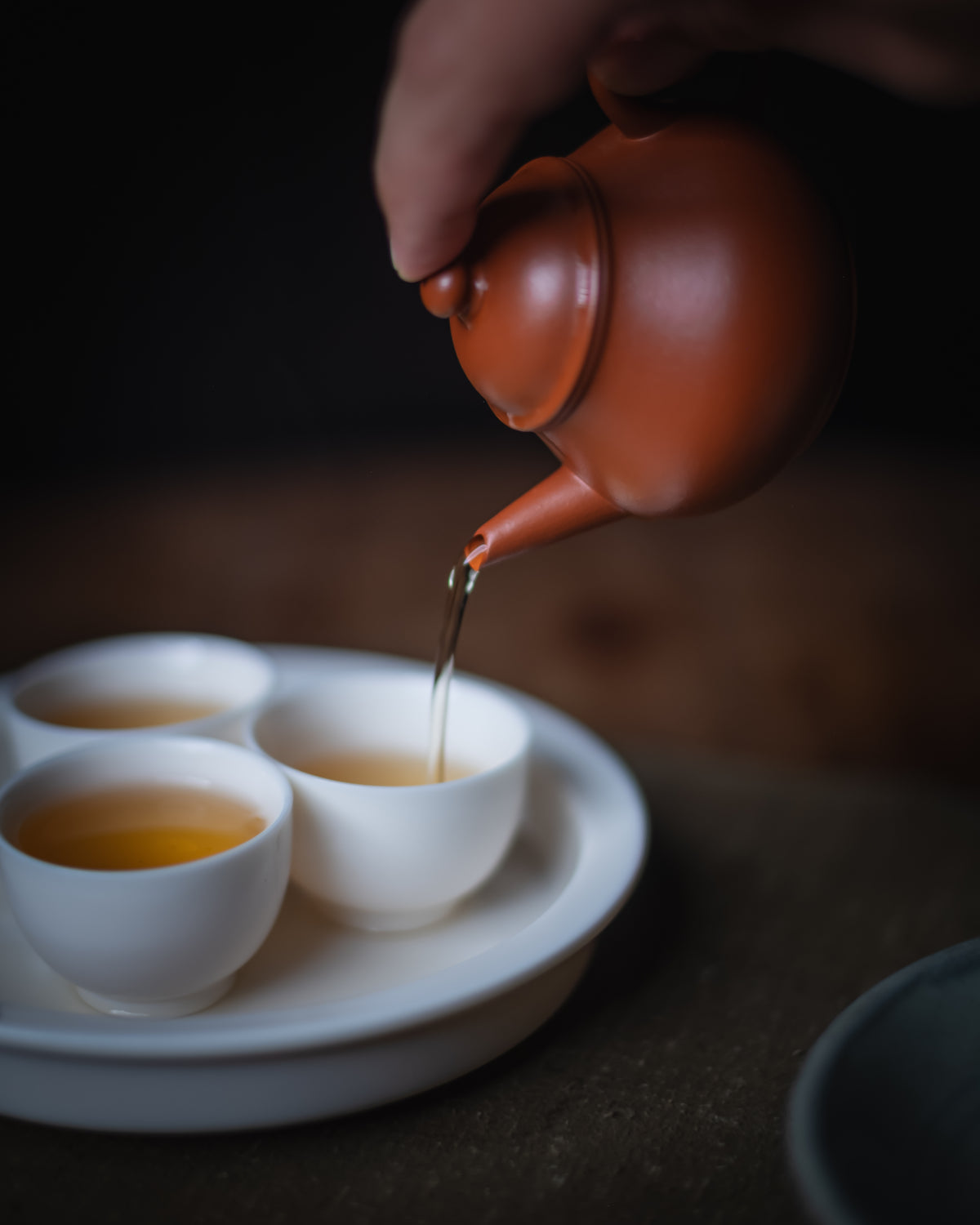 A hand pouring tea from a terracotta clay teapot into three small white cups arranged on a white tray, photographed in a dim, moody setting.