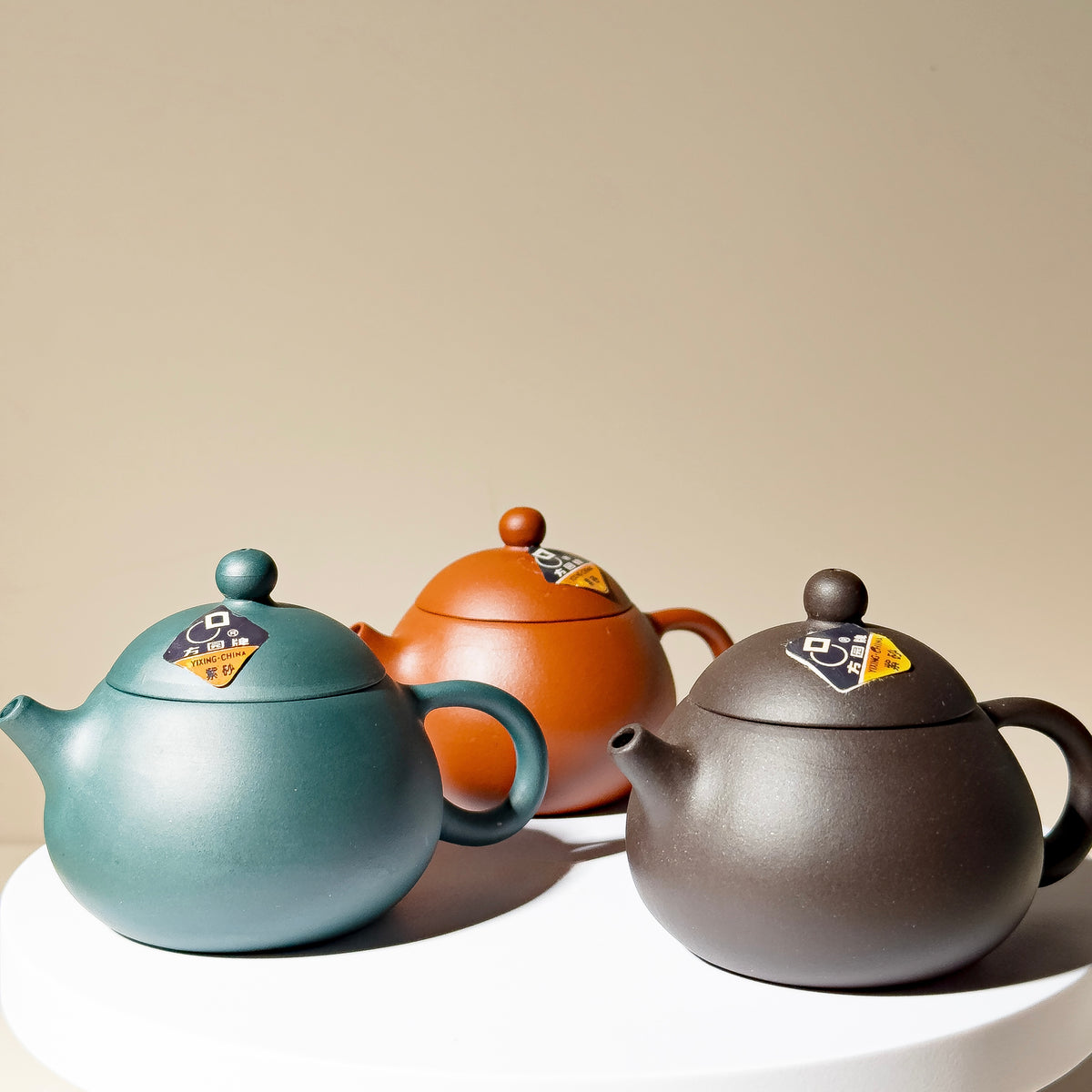 Three ceramic teapots in teal, terracotta orange, and dark charcoal gray, arranged on a white round pedestal against a warm beige background.