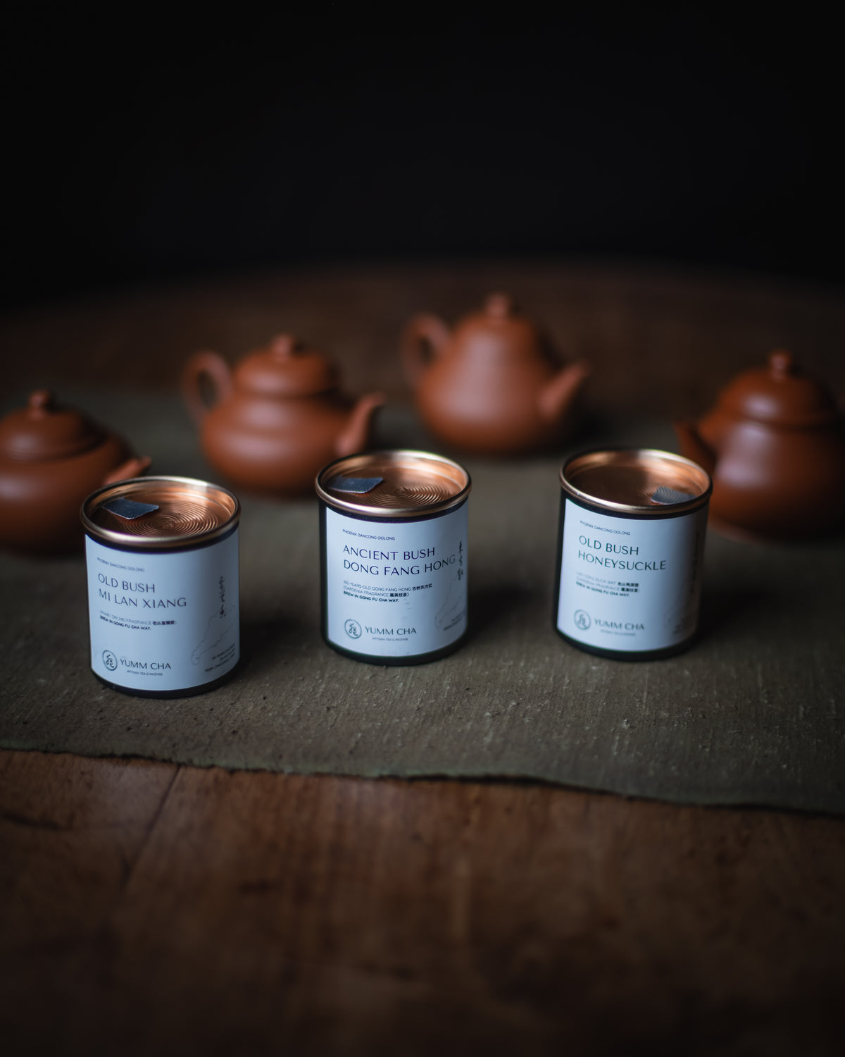 Three tea tins with white labels and metallic lids are arranged on a dark cloth over a wooden surface, with blurred terracotta teapots in the background.
