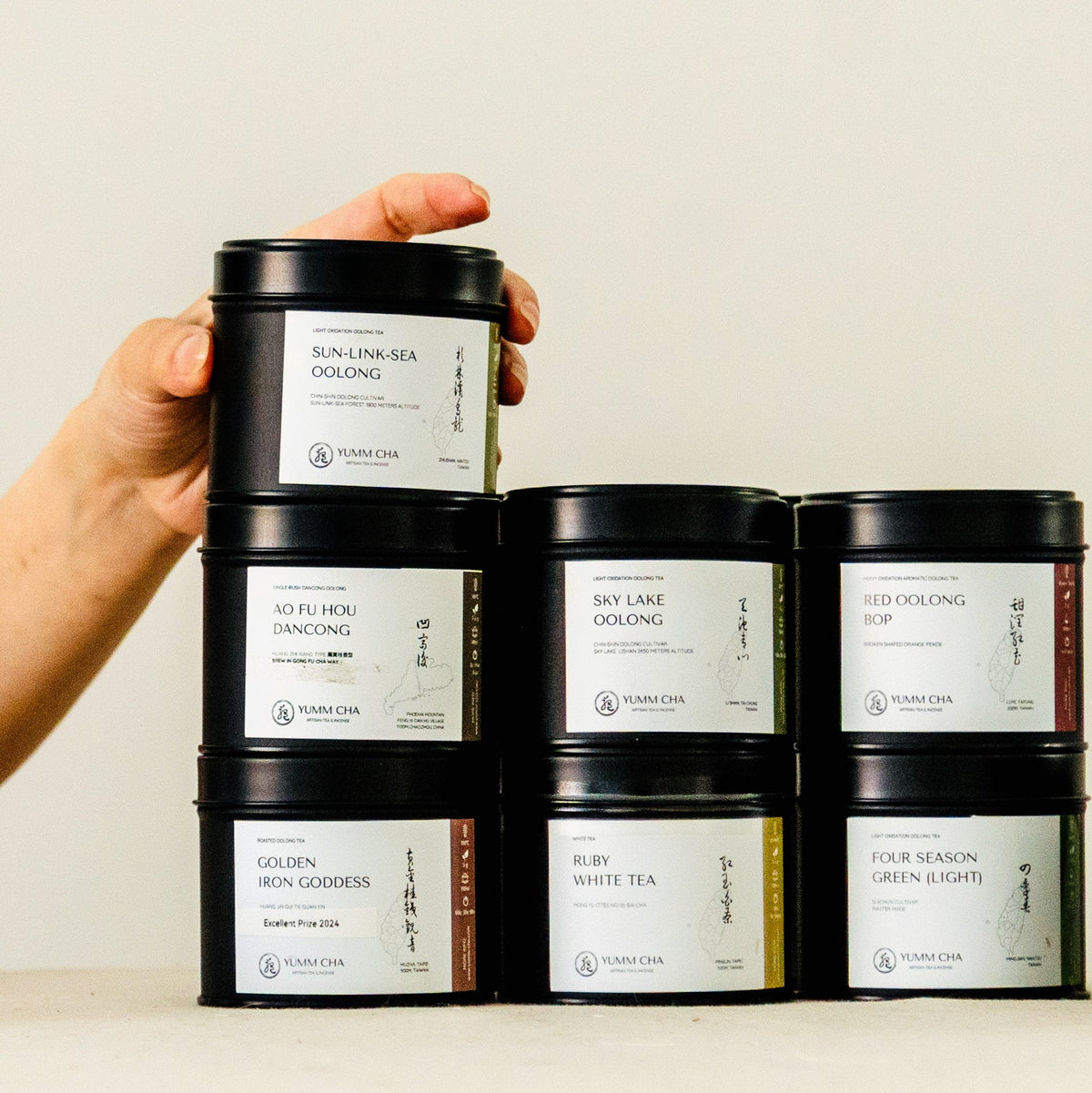 A hand holds a stack of black tea tins against a plain light beige background. The tins have white labels with black text and are arranged in a pyramid with five tins on the bottom level and two stacked on top.